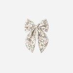 A fabric hair bow accessory featuring a delicate floral print in muted pink, olive green, and brown on a cream background, centered on a light gray studio background.