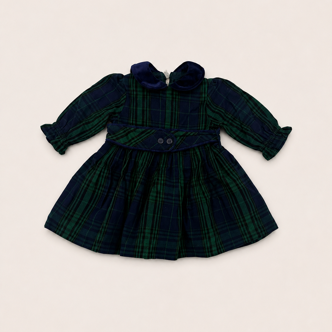 A long-sleeve plaid dress for toddlers, featuring a dark green and navy tartan pattern, a matching waist tie with two buttons, and a ruffled collar, laid flat on a light beige background.