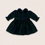 A long-sleeve plaid dress for toddlers, featuring a dark green and navy tartan pattern, a matching waist tie with two buttons, and a ruffled collar, laid flat on a light beige background.