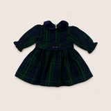 A long-sleeve plaid dress for toddlers, featuring a dark green and navy tartan pattern, a matching waist tie with two buttons, and a ruffled collar, laid flat on a light beige background.