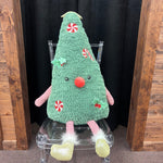 A plush Christmas tree toy with a fuzzy green body, red nose, red-and-white candy cane decorations, pink striped arms and legs, and pale yellow boots, sitting on a clear acrylic chair between wooden panels and a black curtain.