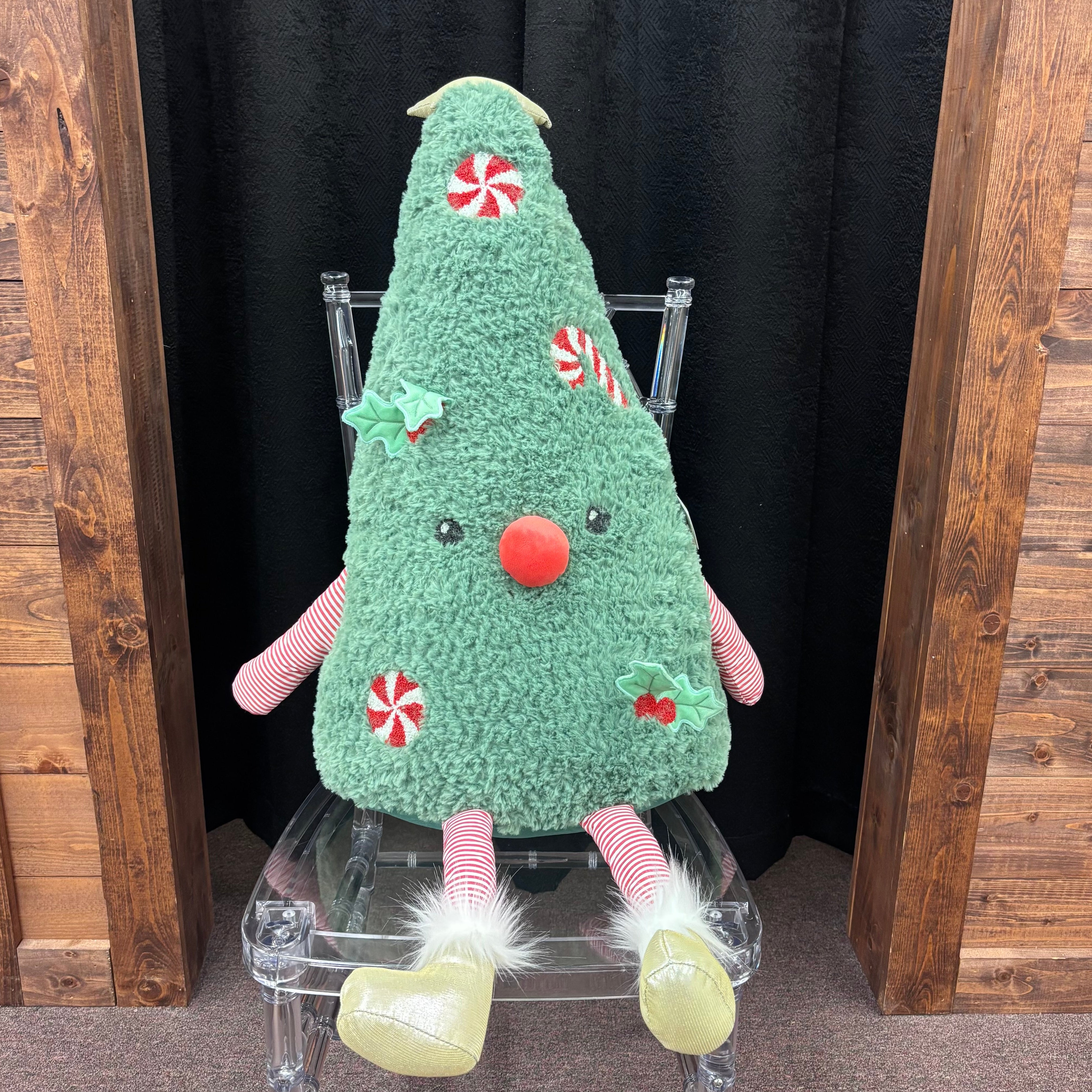 A plush Christmas tree toy with a fuzzy green body, red nose, red-and-white candy cane decorations, pink striped arms and legs, and pale yellow boots, sitting on a clear acrylic chair between wooden panels and a black curtain.
