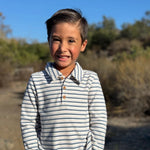A young boy smiling outdoors, wearing a long-sleeve polo shirt with thin navy and off-white horizontal stripes, paired with dark gray jeans.