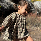 A young child wearing a short-sleeve plaid dress outdoors, standing in a natural setting with rocks and dry grass in the background.