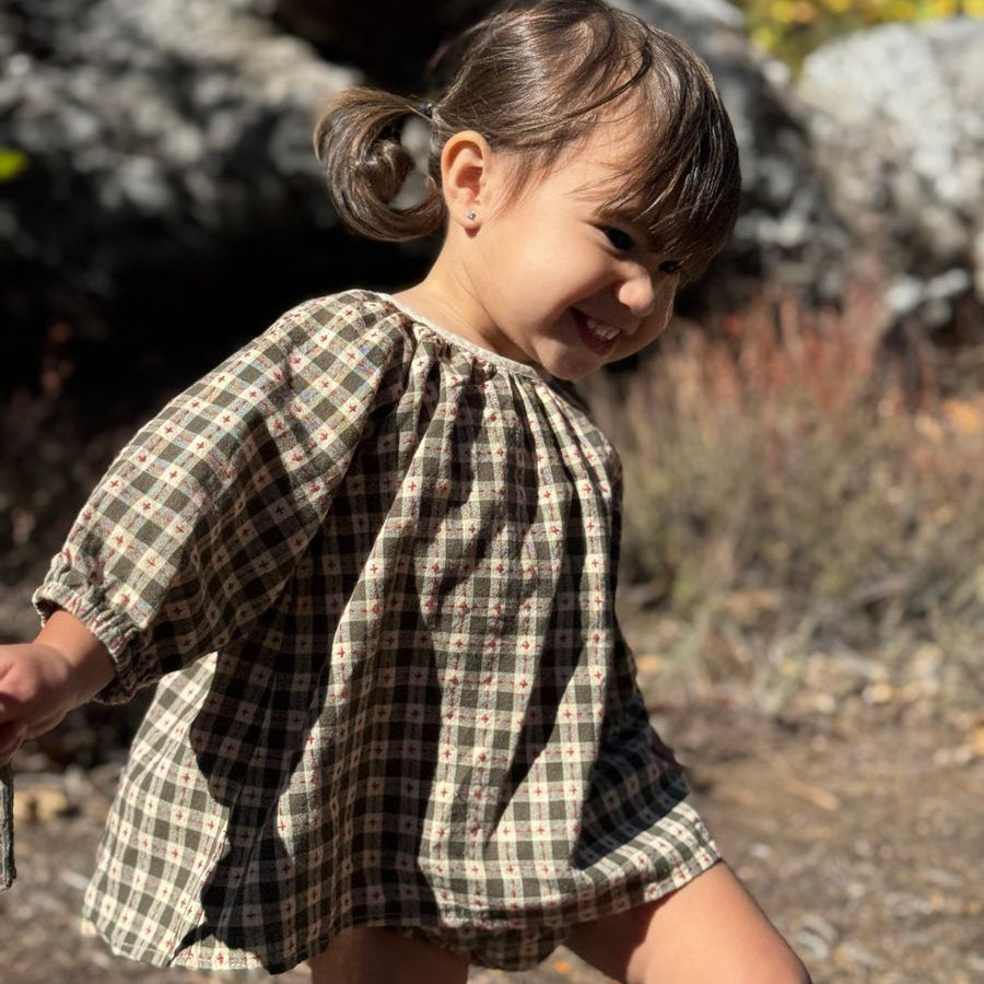 A young child wearing a short-sleeve plaid dress outdoors, standing in a natural setting with rocks and dry grass in the background.