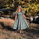 A woman wearing a light sage green tiered midi dress with puff sleeves stands outdoors in a wooded area with dry grass and autumn trees.