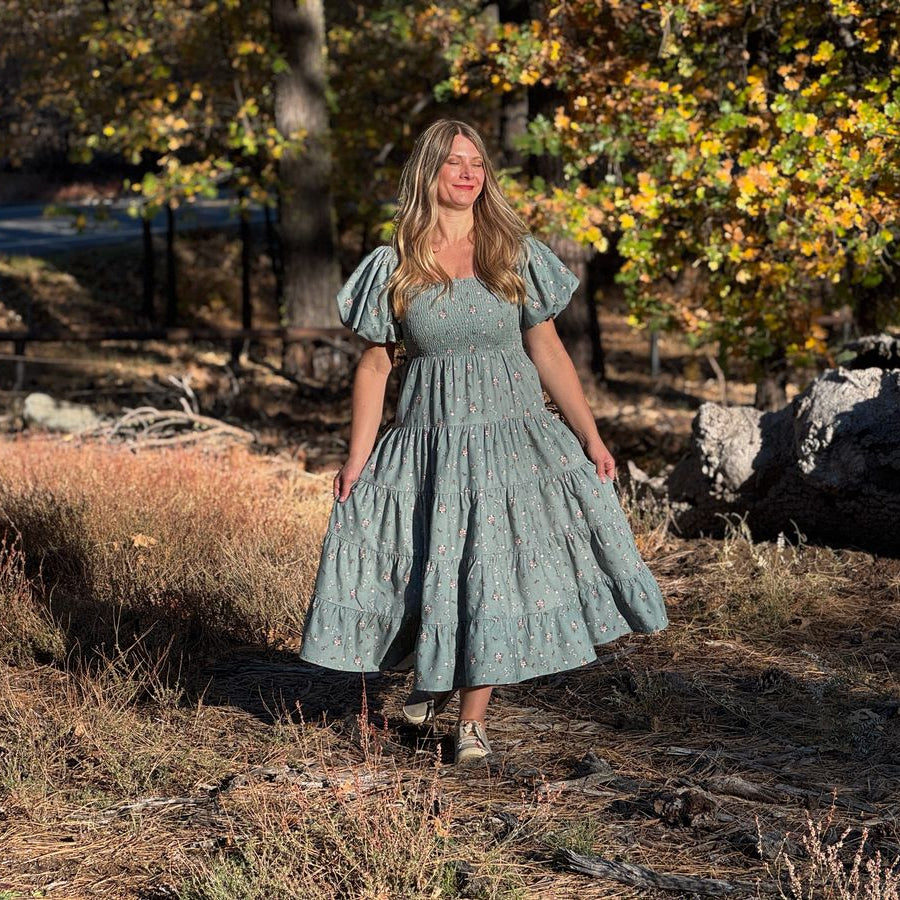 A woman wearing a light sage green tiered midi dress with puff sleeves stands outdoors in a wooded area with dry grass and autumn trees.