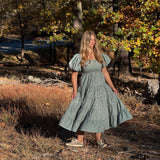 A woman wearing a sage green puff-sleeve midi dress stands outdoors in a wooded area with autumn trees and dry grass.