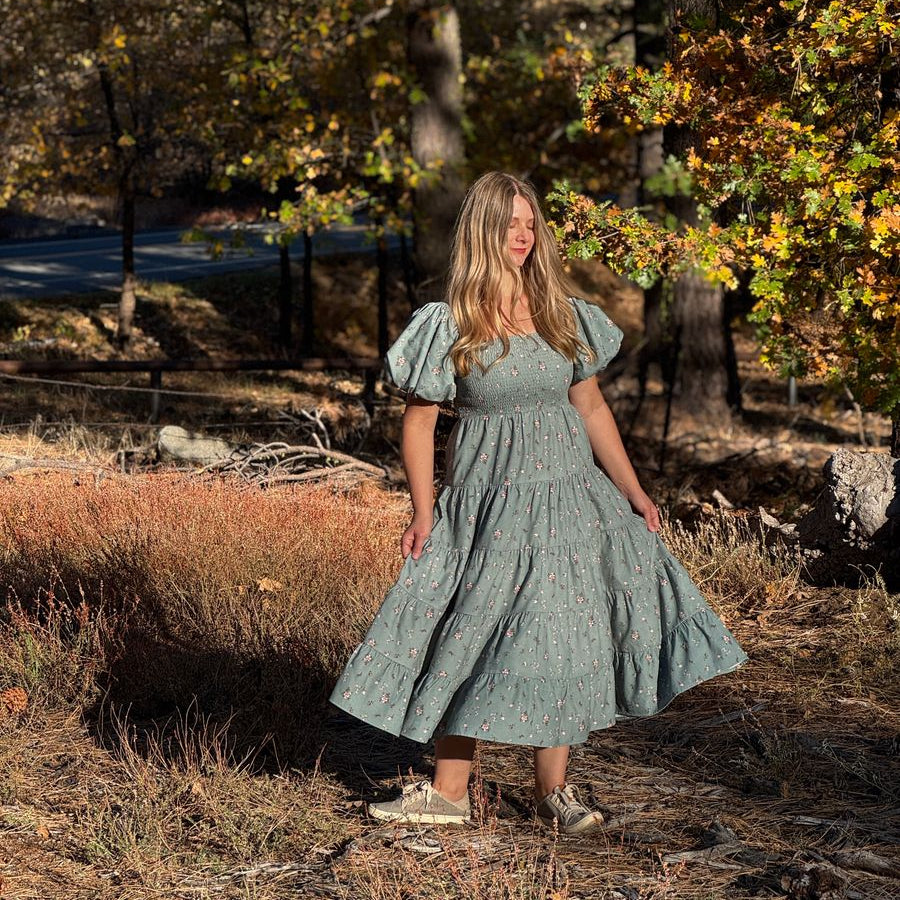A woman wearing a sage green puff-sleeve midi dress stands outdoors in a wooded area with autumn trees and dry grass.