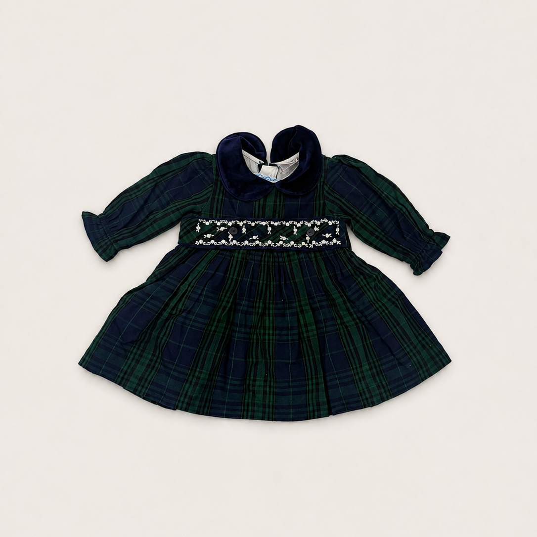 A baby girl's long-sleeve plaid dress in dark green and navy tartan, featuring a smocked bodice with white embroidery and a dark navy heart-shaped collar, laid flat on a light beige background.