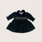 A baby girl's long-sleeve plaid dress in dark green and navy tartan, featuring a smocked bodice with white embroidery and a dark navy heart-shaped collar, laid flat on a light beige background.