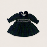 A baby girl's long-sleeve plaid dress in dark green and navy tartan, featuring a smocked bodice with white embroidery and a dark navy heart-shaped collar, laid flat on a light beige background.