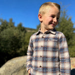 Young boy wearing a plaid shirt standing outdoors with trees and blue sky in the background