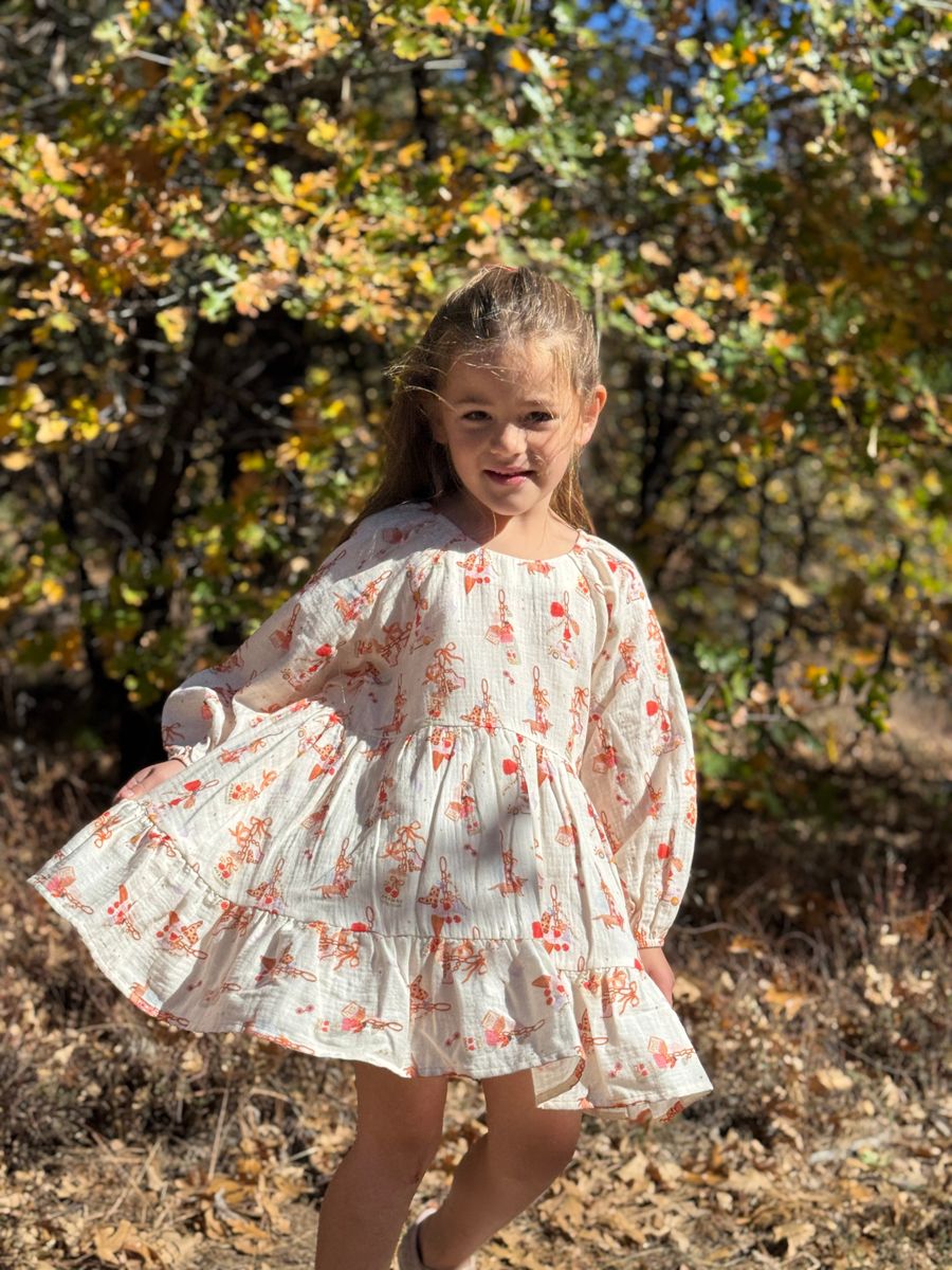 A young girl wearing a white long-sleeve cotton dress with a red and orange floral print, standing outdoors in a wooded area with autumn foliage.