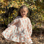 A young girl wearing a white long-sleeve cotton dress with a red and orange floral print, standing outdoors in a wooded area with autumn foliage.