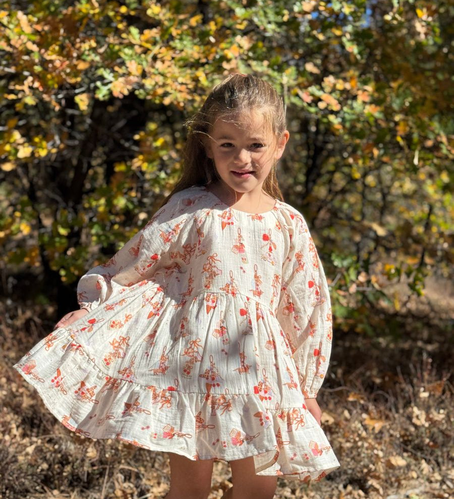 A young girl wearing a white long-sleeve cotton dress with a red and orange floral print, standing outdoors in a wooded area with autumn foliage.