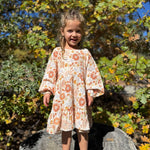 A young girl with blonde hair stands on a tree stump outdoors, wearing a long-sleeve tiered dress with a peach and orange floral pattern on a cream background. The background is filled with green and yellow autumn foliage.
