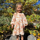 A young girl with blonde hair stands on a tree stump outdoors, wearing a long-sleeve tiered dress with a peach and orange floral pattern on a cream background. The background is filled with green and yellow autumn foliage.