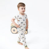 A toddler standing barefoot in a white short-sleeve pajama set with a tractor print, photographed on a clean white studio background with a wooden toy cart behind them.