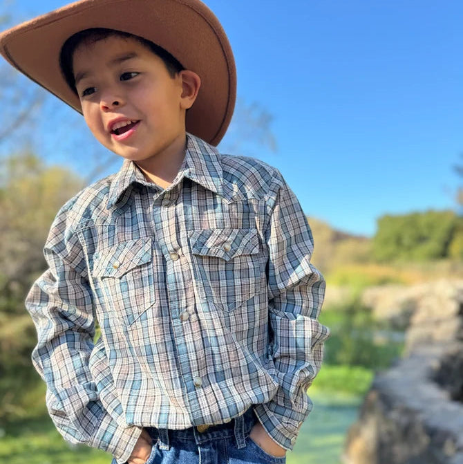 A young boy wearing a brown cowboy hat, a blue and beige plaid western long-sleeve button-up shirt, and blue jeans, standing outdoors against a bright blue sky and blurred green landscape.