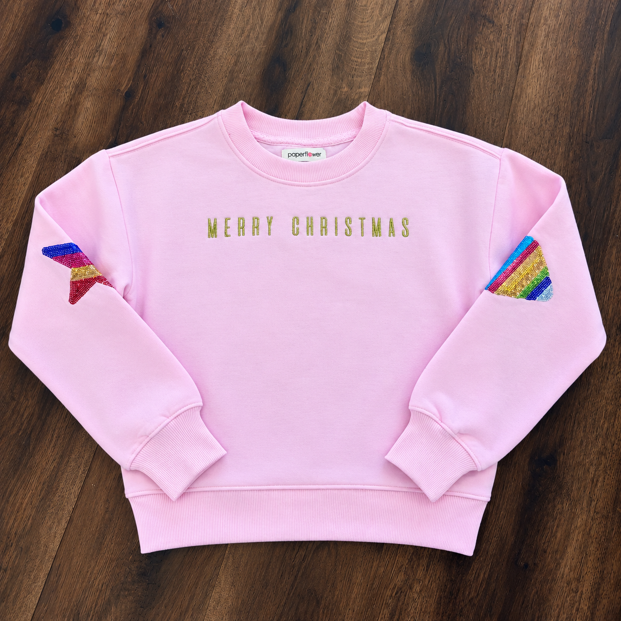 A light pink crewneck sweatshirt with 'MERRY CHRISTMAS' printed in gold across the chest. The sleeves feature pockets containing rainbow-striped fabric pieces, and the sweatshirt is laid flat on a dark wood background.