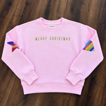 A light pink crewneck sweatshirt with 'MERRY CHRISTMAS' printed in gold across the chest. The sleeves feature pockets containing rainbow-striped fabric pieces, and the sweatshirt is laid flat on a dark wood background.