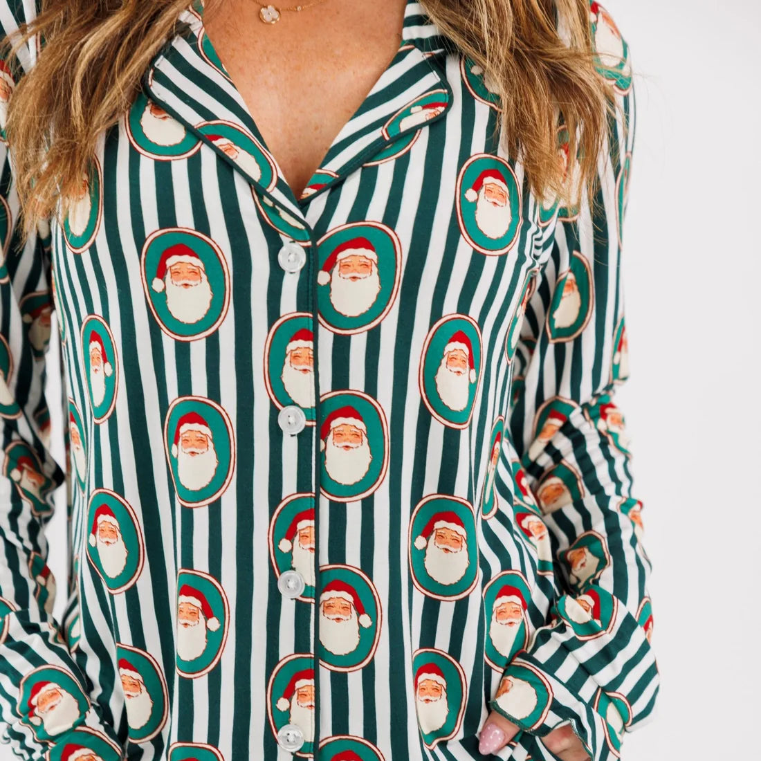 A person wearing a long-sleeve pajama set with dark green and white vertical stripes and an allover Santa Claus face print, shown against a plain light background.