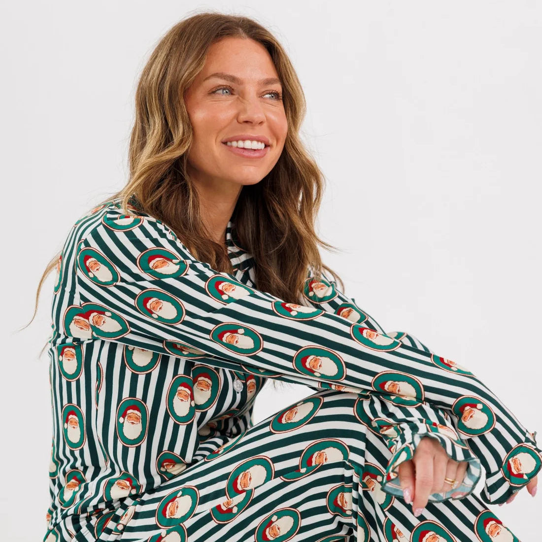 A woman sitting cross-legged wearing a long-sleeve pajama set with dark green and white stripes and a repeating Santa Claus face pattern, photographed against a clean white studio background.