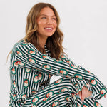 A woman sitting cross-legged wearing a long-sleeve pajama set with dark green and white stripes and a repeating Santa Claus face pattern, photographed against a clean white studio background.