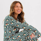 A woman sitting cross-legged wearing a long-sleeve pajama set with dark green and white stripes and a repeating Santa Claus face pattern, photographed against a clean white studio background.