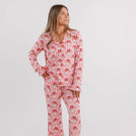 A woman modeling a matching long-sleeve pajama set with a pink and red pattern, standing against a plain light gray studio background.