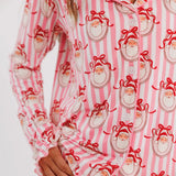 A close-up of a person wearing pink and white striped pajamas with a repeating Santa Claus face print. The pajama top has long sleeves, a button placket, and ruffled cuffs, while the matching pants are also printed.