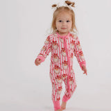 A toddler girl walking barefoot in a pink and white striped one-piece pajama with a cartoon bear print, wearing a small white bow headband, against a plain white studio background.
