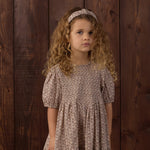 A young girl with curly light brown hair wearing a short-sleeved, tiered dress with a small beige and brown floral pattern, standing against a dark wood plank background.