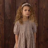 A young girl with curly light brown hair wearing a short-sleeved, tiered dress with a small beige and brown floral pattern, standing against a dark wood plank background.