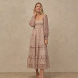 A woman models a long-sleeved midi dress with a tiered ruffle hem and a delicate floral pattern in dusty pink and beige, set against a textured beige studio backdrop.