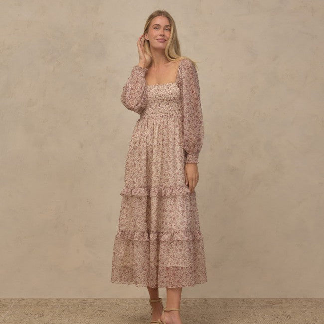 A woman models a long-sleeved midi dress with a tiered ruffle hem and a delicate floral pattern in dusty pink and beige, set against a textured beige studio backdrop.