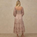 A woman stands with her back to the camera wearing a dusty pink tiered maxi dress with a delicate floral pattern and smocked bodice, paired with beige strappy heels, against a textured beige studio backdrop.
