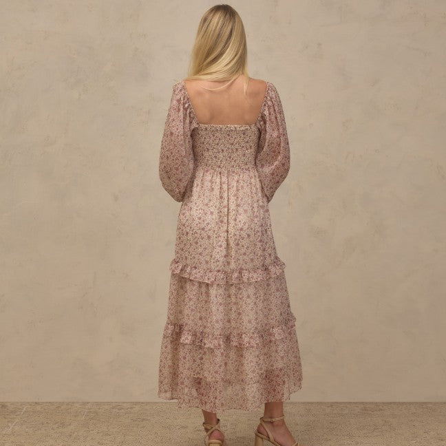 A woman stands with her back to the camera wearing a dusty pink tiered maxi dress with a delicate floral pattern and smocked bodice, paired with beige strappy heels, against a textured beige studio backdrop.