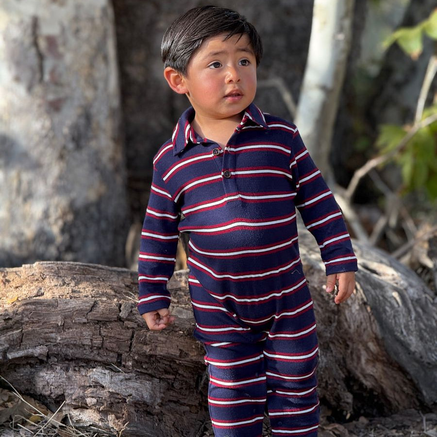 A toddler standing outdoors in front of tree trunks, wearing a navy long-sleeve one-piece outfit with thin red and white horizontal stripes.