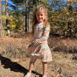 A young girl stands outdoors in a wooded area wearing a long-sleeve striped dress with a gathered skirt and matching shorts. The dress has thin horizontal stripes in peach, pink, orange, green, and white.