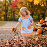 A young girl with blonde hair smiling and playing in a pile of autumn leaves on a path, wearing a light blue dress with orange trim and pumpkin appliques. Pumpkins and a bench are visible in the background with colorful fall trees.
