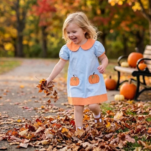 A young girl with blonde hair smiling and playing in a pile of autumn leaves on a path, wearing a light blue dress with orange trim and pumpkin appliques. Pumpkins and a bench are visible in the background with colorful fall trees.