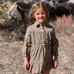 A young girl smiling outdoors in a brown and beige plaid long-sleeve dress with a button-up front and ruffled skirt, standing in a natural wooded setting.