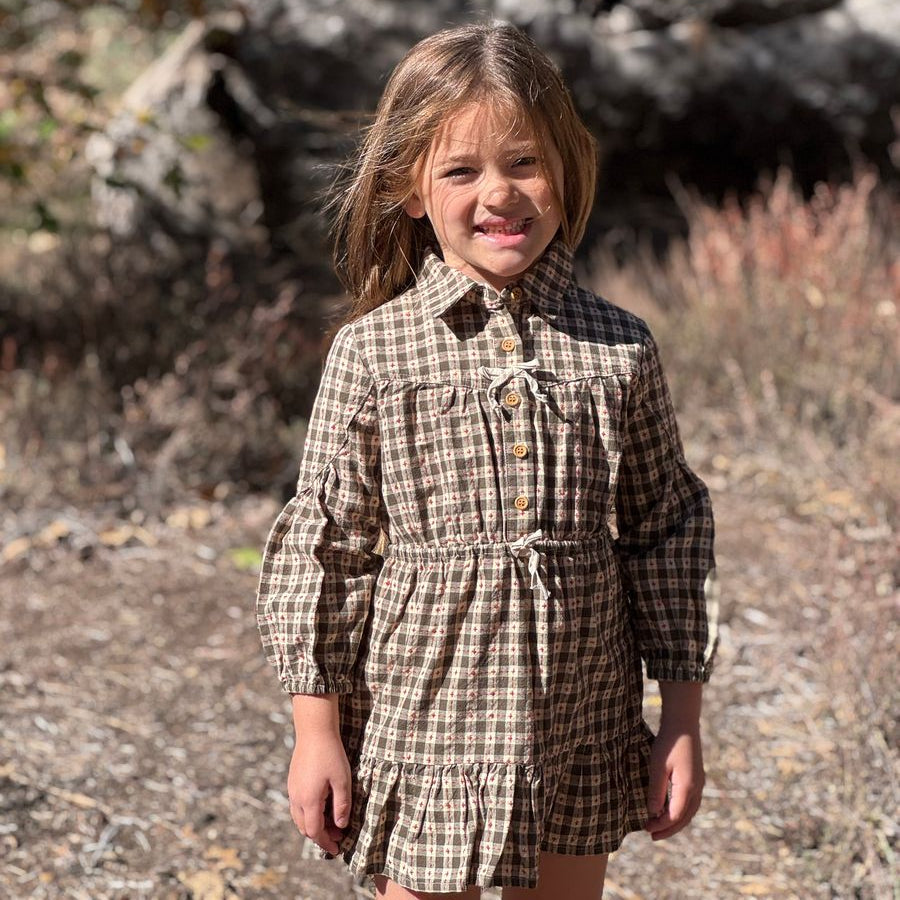 A young girl smiling outdoors in a brown and beige plaid long-sleeve dress with a button-up front and ruffled skirt, standing in a natural wooded setting.