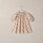 A short-sleeve baby dress with a delicate floral print in muted blue and brown on a soft beige background, hanging on a simple metal hanger against a textured beige wall.