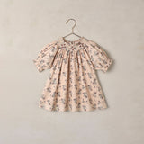 A short-sleeve baby dress with a delicate floral print in muted blue and brown on a soft beige background, hanging on a simple metal hanger against a textured beige wall.