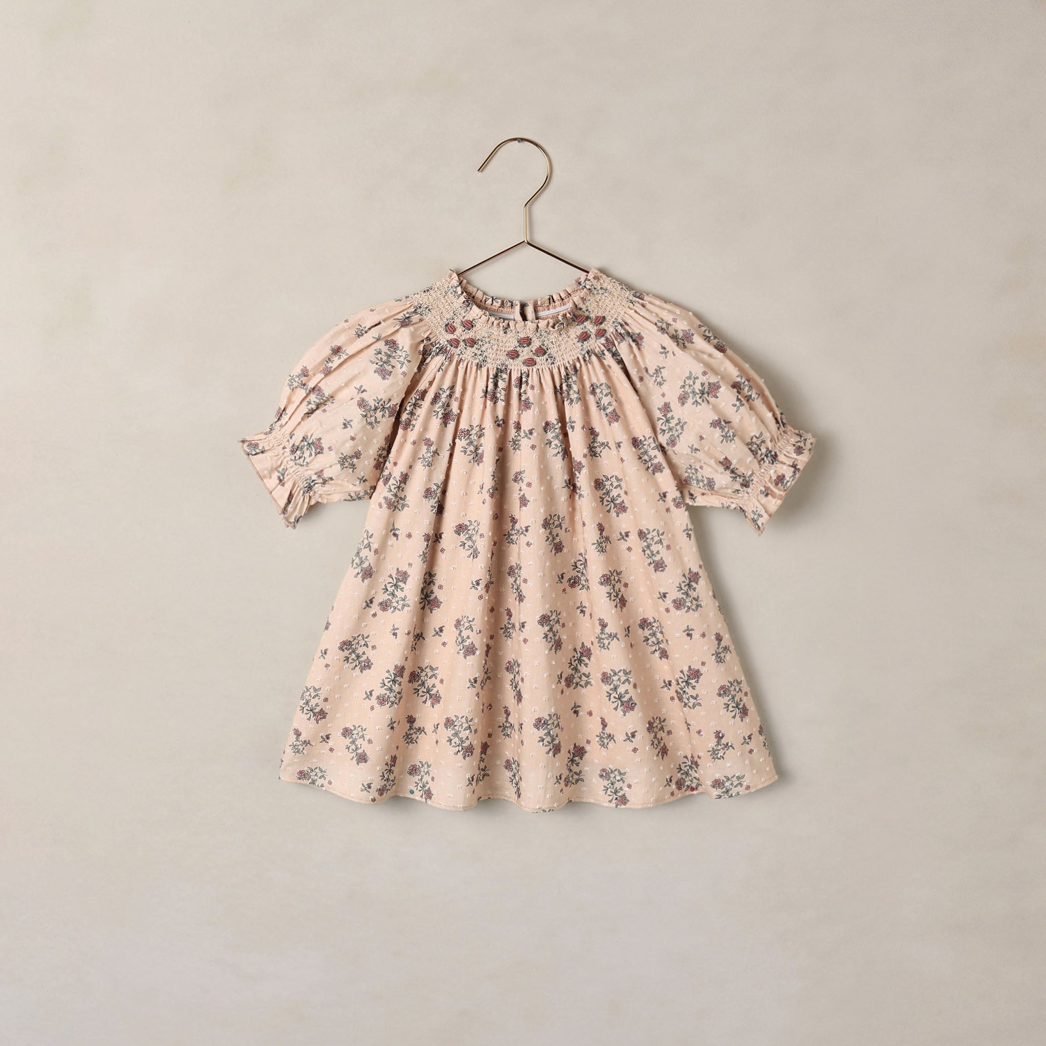 A short-sleeve baby dress with a delicate floral print in muted blue and brown on a soft beige background, hanging on a simple metal hanger against a textured beige wall.