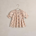 A children's short sleeve dress with a smocked bodice and ruffled sleeves, featuring a delicate floral print in muted blue and green on a petal pink. The dress is shown hanging on a simple metal hanger against a textured, warm beige wall.