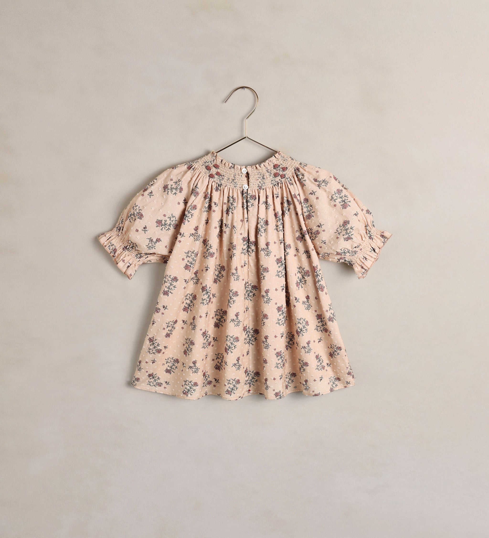 A children's short sleeve dress with a smocked bodice and ruffled sleeves, featuring a delicate floral print in muted blue and green on a petal pink. The dress is shown hanging on a simple metal hanger against a textured, warm beige wall.